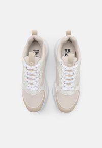 Zapatillas beige y blancas con parte superior de malla, acentos texturizados, cordones planos y suela acolchada. Incluye una asa trasera para facilitar el uso.