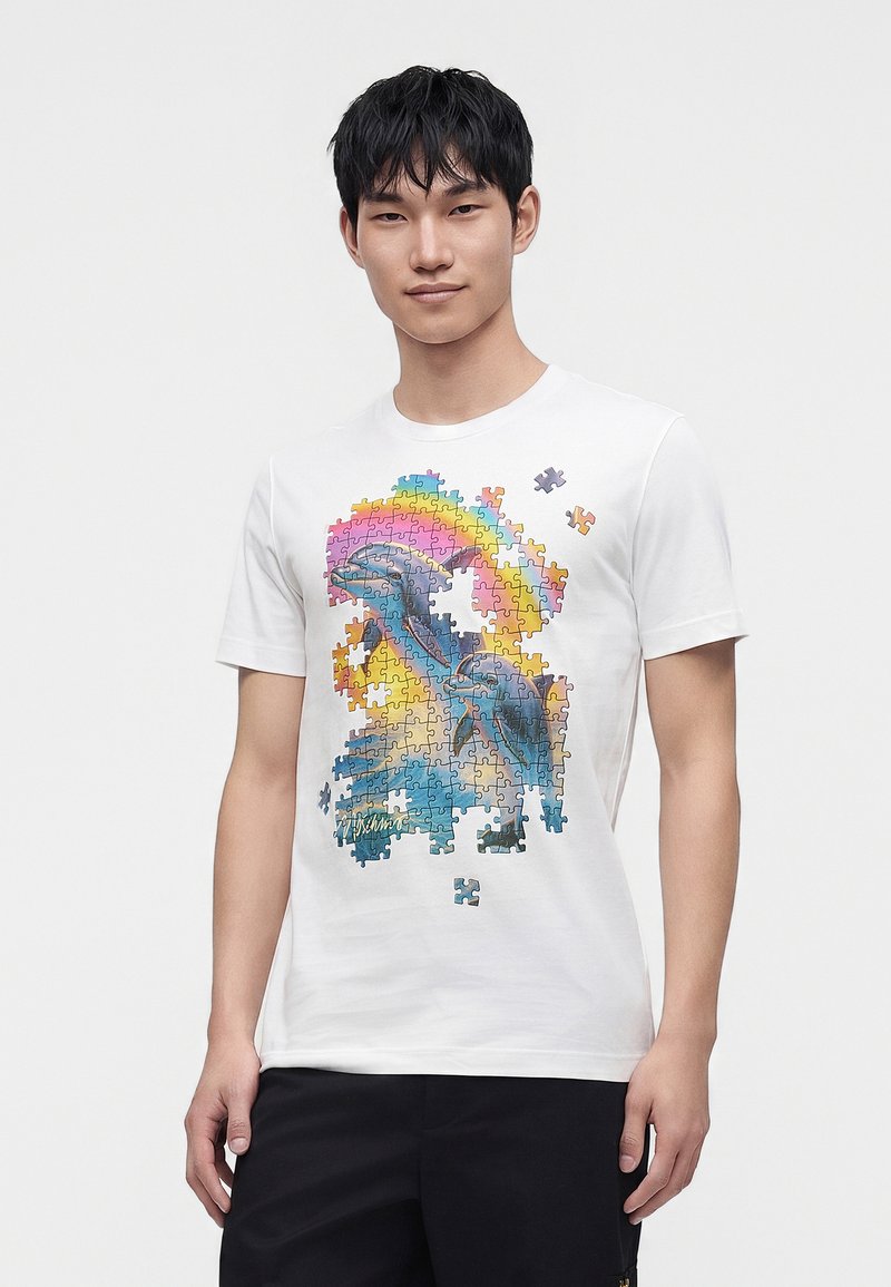 Νέος άνδρας φοράει λευκό T-shirt με πολύχρωμο σχέδιο παζλ δελφινιού, με κάποια κομμάτια του παζλ να λείπουν και να αιωρούνται πάνω από το σχέδιο.