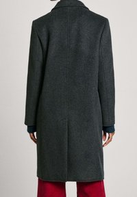 Donkergrijze wollen jas met een getailleerde pasvorm, een nietched kraag en lange mouwen, die tot halverwege de dij reikt. Heeft een strak ontwerp aan de achterkant.