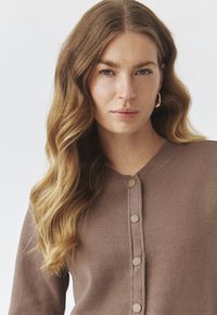 Brauner, gestrickter Cardigan mit Rundhalsausschnitt, der sechs passende Knöpfe auf der Vorderseite und ein geripptes Muster entlang des Kragens und der Bündchen aufweist.