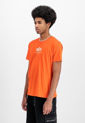 Oranges Baumwoll-T-Shirt mit kurzen Ärmeln, Rundhalsausschnitt und einem weißen Logo auf der Brust. Getragen mit schwarzen Cargo-Shorts, die Taschen haben.
