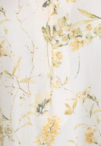 Květinová blůza s žlutým a zeleným botanickým vzorem na krémovém pozadí. Má malé bílé knoflíky na přední části. Měkká textura.