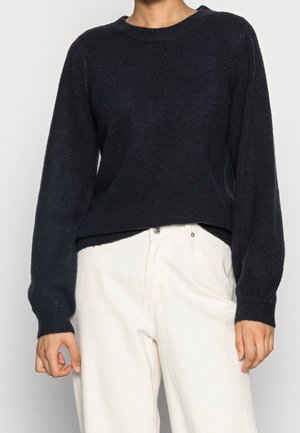 Personne portant un pull noir à manches longues et un pantalon blanc taille haute, debout devant un fond clair uni.