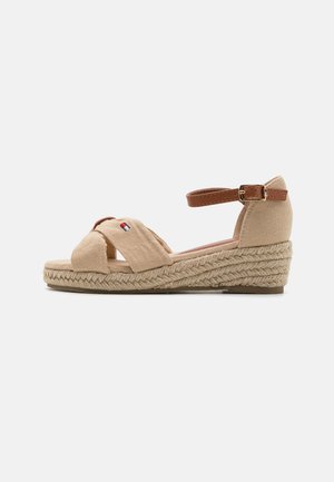 Sandales compensées beiges avec des sangles en tissu croisées, une bride de cheville marron avec une boucle, et une semelle en jute. Semelle extérieure en caoutchouc plate.