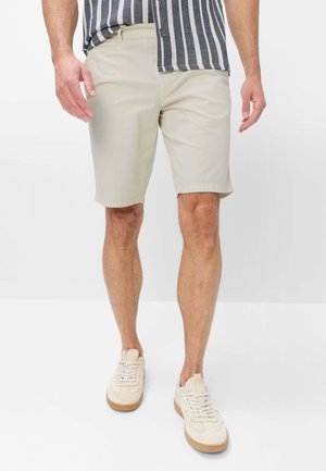 Mann trägt beige knielange Shorts, weiße Sneaker und ein schwarz-weiß gestreiftes Hemd, bei dem eine Seite herausgezogen ist.
