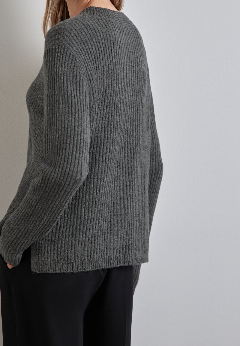Femme portant un pull en maille gris côtelé à manches longues et un pantalon noir, debout devant un mur gris clair uni.