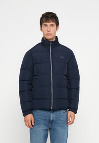 Young man wearing a navy blue quilted jacket with front zipper and blue jeans, standing against a plain white background.