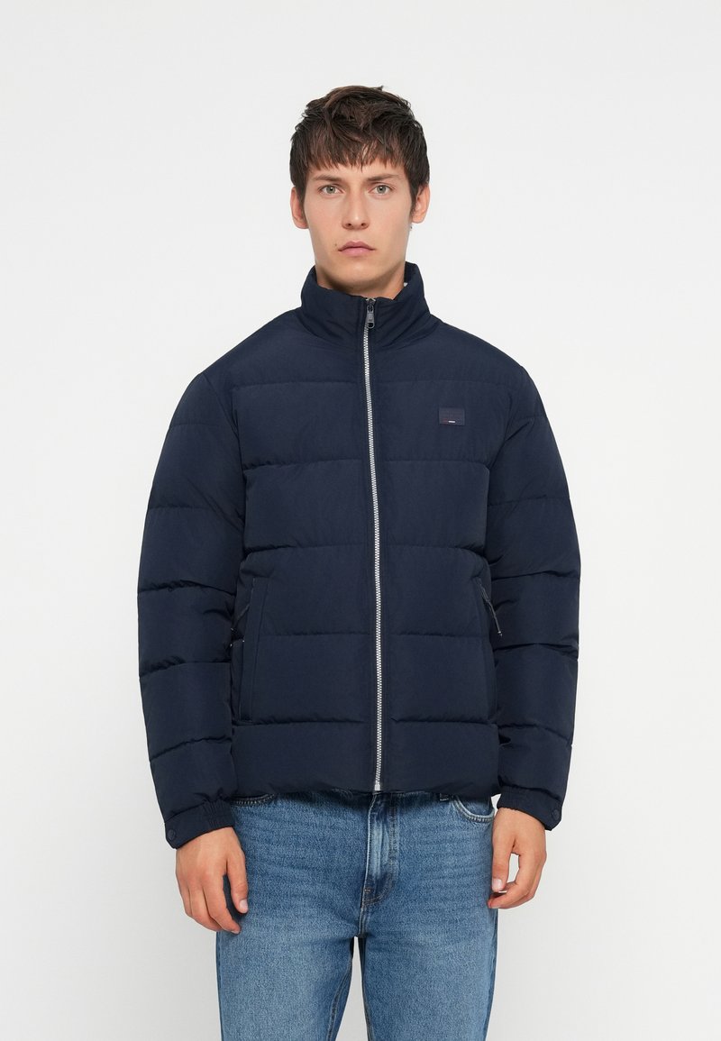 Young man wearing a navy blue quilted jacket with front zipper and blue jeans, standing against a plain white background.