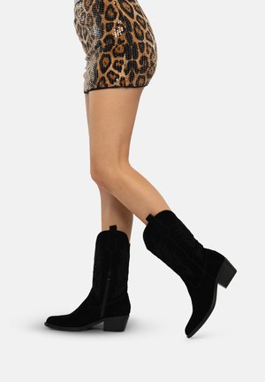 Stivali da cowboy neri in suede con punta affilata, dettagli ricamati sui lati e tacco corto impilato, abbinati a una gonna con stampa leopardo.