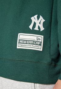 Grønn hettegenser med hvit brodert NY-logo og et rektangulært hvitt merke med påskriften "NEW ERA CAP" i svart tekst nær livet.