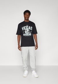 Juodas grafikos marškinėliai su "Las Vegas Raiders" tekstu ir amerikietiško futbolo šalmą dizainu. Suderinta su šviesiai pilkais sportiniais kelnais ir baltais sportbačiais.