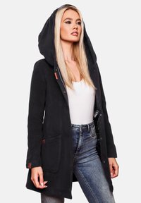 Manteau à capuche noir en matière douce, avec des poches devant, une fermeture éclair et des accents de boutons. Porté sur un haut blanc et un jean.