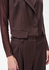 Brązowa, dopasowana kamizelka z usztywnianym kołnierzem, podwójnym zapięciem na guziki i bocznymi kieszeniami, zestawiona z pasującymi, wysokim stanem spodniami.