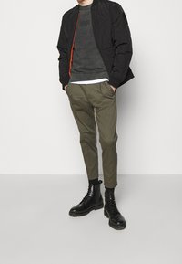 Czarna pikowana kurtka bomberka z pomarańczowym zamkiem błyskawicznym, szara bluza, oliwkowe spodnie zwężane oraz czarne sznurowane botki. Gładkie tekstury na całej powierzchni.