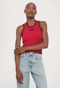 Raudona lygi tankų palaidinė su juodais akcentais, su "Moschino Jeans" logotipu. Suderinta su šviesiai mėlynomis džinsais, sukuriant atsitiktinį stilių.