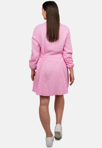 Abito rosa con texture, maniche lunghe e vita legata, caratterizzato da una silhouette svasata e un orlo a ginocchio. Indossato con sneakers bianche.