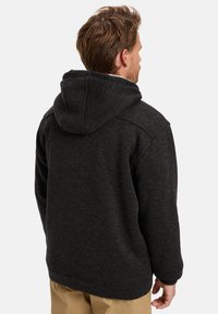 Uomo con capelli castani chiari che indossa una felpa nera con cappuccio e pantaloni beige, visto da dietro su uno sfondo semplice.