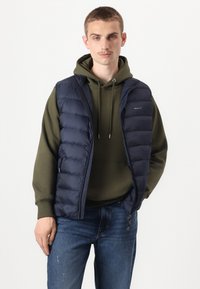 Mornarska puffer prsluk sa quilts dizajnom, prednjim zatvaračem i bočnim džepovima, nosi se preko maslinasto zelene dukserice s kapuljačom. Opušteni kroj, lagani materijal.