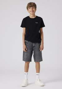 Černé tričko s krátkým rukávem a logem, šedé džínové šortky, bílé ponožky a bílé tenisky, stojící postoj na světle šedém pozadí.