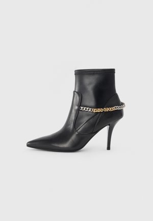 Stivaletto in pelle nera con punta affusolata, alto tacco a stiletto e decorazione a catena dorata e argento attorno alla caviglia.