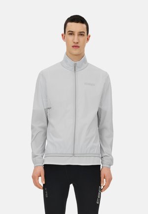 Young man wearing a light gray zip-up jacket and black pants with sport branding, standing against a plain white background.