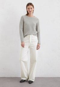 Lange grijze top met lange mouwen van zacht materiaal, gecombineerd met witte wijde jeans met een knoopsluiting en zakken, afgewerkt met zwarte puntige schoenen.