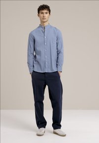 Svetlo modra srajca z gumbi in pokončnim ovratnikom, izdelana iz bombaža, v kombinaciji z mornarsko modrimi hlačami. Gladka tekstura, kroj prilagojen postavi. Bele superge.