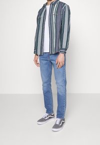 Camisa de botones a rayas verdes y blancas sobre una camiseta blanca, combinada con jeans de color azul claro y zapatillas grises.