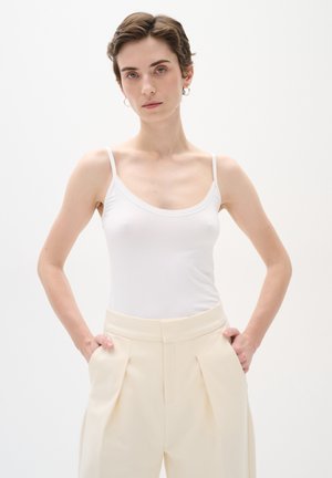 Haut sans manches blanc avec un col rond, associé à un pantalon beige taille haute avec des plis devant et des poches latérales.