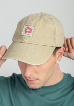 Uomo che si sistema un berretto da baseball beige con toppa floreale rosa e gialla con la scritta "Kaotiko Society", indossa una maglietta turchese e un orecchino a bottone singolo.