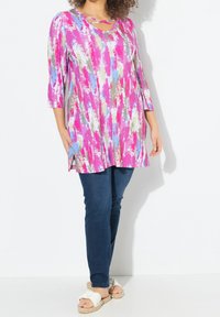 Mehrfarbige Tunika mit abstrakten Mustern in Rosa, Blau und Grau; mit einem Rundhalsausschnitt und Kreuzträgern sowie Dreiviertelärmeln. Kombiniert mit dunklen Jeans und hellen Sandalen.