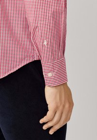 Bras gauche et main portant une chemise à manches longues à carreaux rouges et blancs avec un poignet boutonné, reposant à côté de pantalons côtelés foncés.