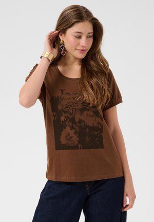 Jeune femme aux longs cheveux bruns portant un t-shirt graphique marron, un jean foncé, un bracelet en or et des boucles d'oreilles pendantes colorées.