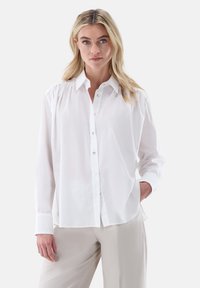 Camicia bianca con bottoni e colletto, caratterizzata da maniche lunghe e una vestibilità morbida. Tessuto liscio con una leggera lucentezza e semplici dettagli con bottoni.