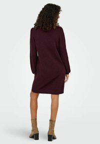 Abito lavorato a maglia bordeaux con maniche lunghe e silhouette dritta, con polsini e orlo a costine. Indossato con stivaletti alla caviglia color taupe.
