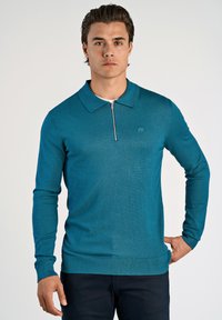 Tealfärgad stickad pullover med dragkedja vid kragen, långärmad och med ribbad fåll. Diskret logotypdetalj på vänster bröst.