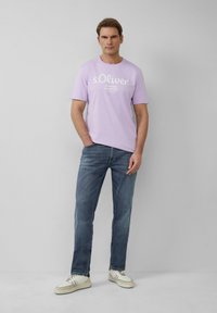 Jasnofioletowa bawełniana koszulka z białym napisem "s.Oliver Elevated Essentials". Połączona z niebieskimi dżinsami i białymi trampkami z brązowymi akcentami.
