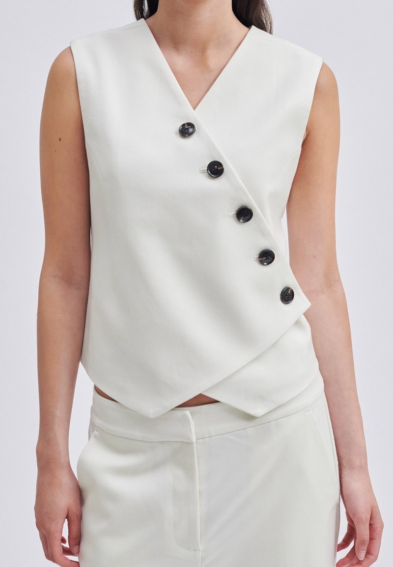 Haut blanc sans manches avec un col en V et un design asymétrique ; comporte cinq boutons noirs le long du devant ; texture de tissu lisse.