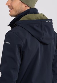 Marineblauwe waterdichte jas met een groene fleece voering, verstelbare capuchon en subtiele logo op de schouder, met een gladde textuur.