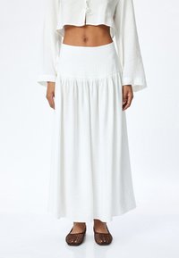 Jupe longue blanche flottante en tissu léger, avec une taille texturée et des plis froncés, associée à des chaussures ouvertes à talons bruns.