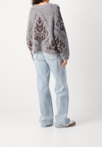 Žena oblečená v šedém a vínovém vzorovaném pleteném svetru, světle modrých širokých džínách a bílých teniskách s gumovými podrážkami, stojící na pozadí bílé stěny.