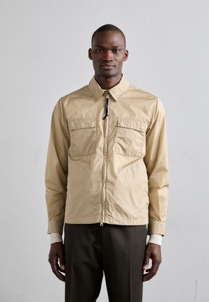 Giacca leggera color beige con zip frontale, colletto a punte, due tasche sul petto, polsini a coste e dettagli a pannello sottili. Indossata sopra pantaloni scuri.