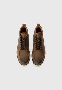 Botas de cuero marrón hasta el tobillo con puntera redonda, acentos en un marrón más claro, cordones negros y un asa en el talón.