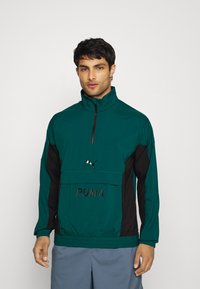 Chaqueta cortavientos en teal y negro con un alto cuello, media cremallera frontal y el logo de "PUMA". Fabricada con un tejido suave y puños elásticos.