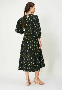 Musta mekko vihreällä kukkakuviolla, puffihihat, polviin ulottuva, avaimenreikäselkäyksityiskohta, yhdistettynä nuden värisiin, hihnallisiiin, lohkolonkkaisiin sandaaleihin.