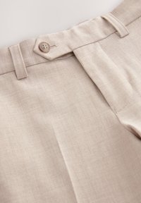 Pantalones beige a medida hechos de tela suave, con cierre de botón, trabillas para cinturón y pliegues marcados. Textura sutil visible.