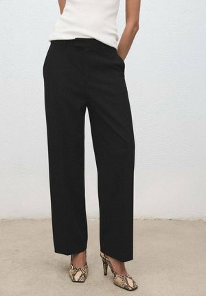 Mujer vestida con pantalones anchos negros, top sin mangas blanco y zapatos de tacón alto con estampado de serpiente, de pie frente a una pared lisa.
