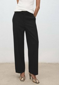 Donna che indossa pantaloni neri a gamba larga, top bianco senza maniche e scarpe con tacco alto a stampa serpente, in piedi contro un muro semplice.