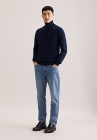 Pull à col roulé bleu marine en tissu fin, associé à un jean bleu clair et des chaussures en cuir noir, se tenant devant un fond neutre.