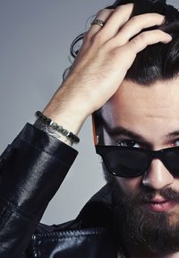 Juodas odinis švarkas, tamsūs akiniai nuo saulės, ranka per plaukus, ant rankos karoliukų apyrankė su pakaitiniais juodais ir pilkais akmenimis.